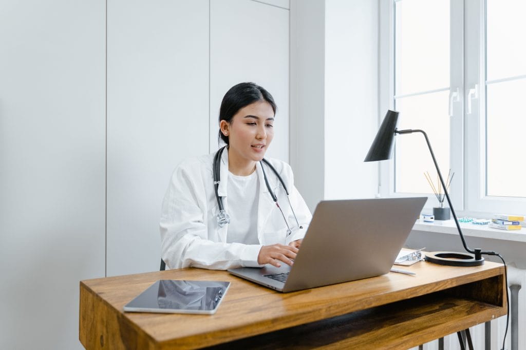 Female doctor taking an English lesson online after her shift. A female doctor in a white coat uses a laptop for an online English lesson from her office during her lunch break.